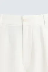 Pantalón blanco de corte relajado (relaxed fit) con pliegues frontales, bolsillos laterales y bolsillos traseros de vivo. Cierre frontal con cremallera y botón.