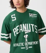 Remera de corte boxy y estilo deportivo, color verde oscuro, con diseño inspirado en el fútbol americano de Peanuts. Presenta cuello redondo, mangas cortas con franjas blancas horizontales y estampas localizadas de Snoopy en el frente.