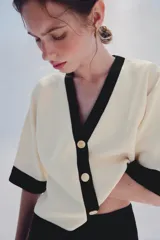 Chaqueta de punto color crudo con ribetes negros en el cuello en V, la botonera, el bajo y los puños de las mangas cortas. Cuenta con cierre frontal con botones dorados grabados.