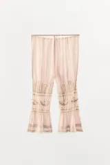 Pantalón de tiro medio confeccionado en viscosa, con cintura elástica ajustable mediante lazada. Presenta detalles de pliegues, bordados florales a tono y aplicaciones de encaje a contraste en la parte inferior, finalizando con un bajo acabado en volantes.