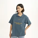 Remera tipo polo de manga corta, color verde oscuro, con cuello y abertura frontal. Presenta un estampado dorado en el pecho con el texto "PulauCrew" y un pequeño logo de corona de laurel bordado.
