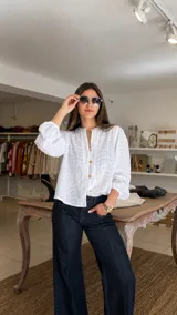 Camisa blanca de broderie con cuello redondo, cierre frontal con botones de madera y mangas largas abullonadas con puños elásticos.