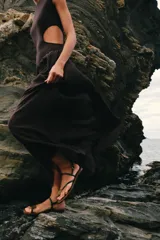 Vestido midi de color negro, sin mangas, con cuello redondo y aberturas laterales en la cintura (cut out). La parte superior es de punto liso y la falda presenta una textura plisada y ligera.