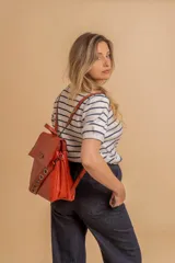 Mochila tipo cartera con tapa, confeccionada en símil cuero color suela. Cuenta con doble compartimento, división interna, cierre de seguridad y bolsillo trasero con cierre. Incluye una correa frontal decorativa con tachas metálicas.