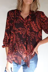 Camisa de lino con estampado paisley en tonos marrón y naranja, cuello con lazos integrados, mangas largas con frunces en hombros y puños elásticos, y cierre frontal con botones de coco.
