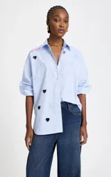 Camisa de algodón a rayas celestes y blancas, con cuello clásico, cierre frontal de botones, mangas largas y aberturas troqueladas en forma de corazón junto a bordados decorativos y texto en el hombro.