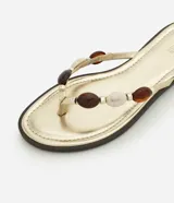 Sandalias planas tipo mule de dedo de la marca Satinato, en color dorado con plantilla metalizada y suela baja. Presenta una tira con aplicaciones decorativas de cuentas de resina en tonos marrones y marfil.