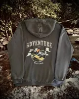 Hoodie oversize color gris oscuro con estampa en la espalda que presenta un paisaje montañoso, árboles y un sol, acompañado por el texto Adventure and Nature.