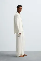Pantalón de corte relajado confeccionado en tejido de cáñamo color blanco roto. Presenta cintura elástica con cordón interior ajustable, cierre frontal con botones, bolsillos laterales y un bolsillo trasero de plastrón.