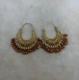 Aros tipo argolla dorados con diseño calado de filigrana y colgantes de pequeñas cuentas rojas en el borde inferior.