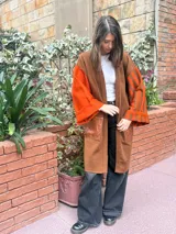 Saco largo de lana merino en color marrón, con diseño abierto sin botones. Presenta mangas anchas con estampado de rayas verticales en tonos naranja y marrón, y un bolsillo frontal aplicado en material símil cuero con textura.
