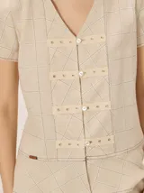 Camisa de manga corta color beige con estampado a cuadros, cuello en V y detalles de galones decorativos estilo oriental en la parte frontal.