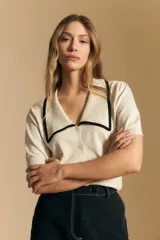 Top de tejido color beige claro, manga corta, escote en V con cierre frontal de botones y ribetes negros en el cuello y el borde del escote.