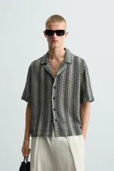 Camisa de calce relajado confeccionada en tejido jacquard con estructura abierta y diseño geométrico. Presenta cuello bowling, manga corta, aberturas laterales en el bajo y cierre frontal con botones.