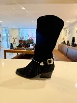 Bota de caña alta estilo western confeccionada en cuero vacuno color suela. Presenta un diseño versátil con estribo desmontable y flecos decorativos en la zona del tobillo, taco de madera y punta fina.