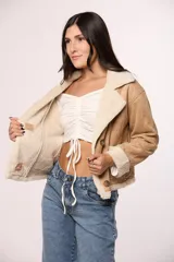 Campera tipo gamulán de gamuza color tostado con interior y solapas de corderito sintético color crema. Presenta cierre frontal con botones, bolsillos cuadrados delanteros y detalles de costuras visibles.
