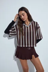 Remera de estilo deportivo con cuello en V y mangas cortas amplias. Presenta un diseño de rayas verticales en tonos marrones, blancos y líneas finas en fucsia, confeccionada en tejido de red.