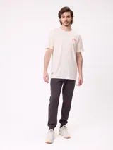 Remera de manga corta color crema con estampado gráfico en la espalda que representa un paisaje de montaña con un lago, árboles y una carpa, junto con el logo de la marca Rusty.