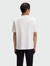 Remera de corte regular, color blanco, con cuello redondo y detalle de logo metálico circular en el pecho.