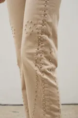 Pantalón de denim negro con diseño de tachas metálicas distribuidas en patrones curvos. Presenta un corte recto y tiro medio.