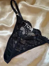 Tanga negra de encaje con diseño de tiras elásticas ajustables y dije con la palabra "TUYA" en pedrería plateada.