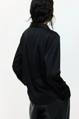 Blusa negra de tela satinada con brillo, cuello clásico, escote en V con botones frontales, mangas largas con puños sencillos y bajo redondeado.