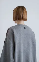 Buzo de cuello redondo con diseño oversize y corte crop. Confeccionado en frisa de algodón color gris melange, presenta un recorte asimétrico en color negro en microfibra con un bolsillo frontal funcional con cierre a presión.