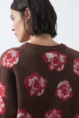 Cardigan de tejido jacquard color marrón con estampado floral en tonos rojo y rosado. Presenta cuello redondo, cierre frontal con botones y terminaciones acanaladas en puños y ruedo.