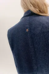 Tapado largo de jean azul oscuro, con cuello solapa y puños con detalle de cinta de terciopelo negro. Presenta costuras visibles que definen paneles y un logo bordado en la espalda.