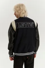 Campera estilo varsity con cuerpo de lana azul marino y mangas de cuero sintético en color negro. Presenta cuello, puños y ruedo acanalados con rayas blancas, cierre frontal con botones a presión y bolsillos laterales.