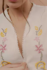 Chaleco color crudo sin mangas, con escote en V y bordados artesanales florales en tono amarillo, rosa y marrón en el frente. Presenta detalles de pasamanería con pompones pequeños en los bordes.