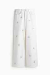 Pantalón de jean blanco, tiro superalto, con piernas amplias y corte holgado. Presenta tachas decorativas en forma de estrella o copo de nieve distribuidas por toda la prenda.