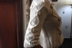 Sweater de lana merino color blanco, de tejido texturado con trenzas y bodoques, cuello redondo y mangas 3/4.