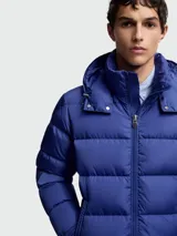 Campera puffer Moncler modelo Maya, color azul, con capucha integrada, cierre frontal bidireccional y bolsillo con solapa en la manga izquierda.