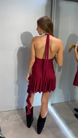 Vestido corto color bordó con acabado brillante, escote tipo cowl neck y espalda descubierta. Presenta un diseño ajustado en el torso con una falda con volados y un detalle de caída lateral asimétrica.