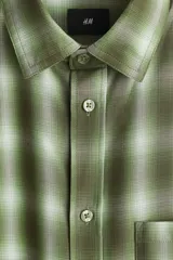 Camisa de manga larga de corte relajado, confeccionada en mezcla de algodón suave con estampado a cuadros en tonos verdes y blancos. Presenta cuello inglés, cierre frontal de botones, un bolsillo aplicado en el pecho y dobladillo redondeado.