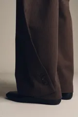 Pantalón de tiro medio color marrón oscuro con textura acanalada o plisada verticalmente. Presenta pierna ancha y cintura con trabillas.