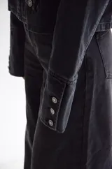 Chaqueta de estilo militar confeccionada en denim negro con efecto lavado. Presenta un diseño estructurado con cierre frontal de cremallera, cuello mao y múltiples botones metálicos decorativos dispuestos en filas horizontales. Incluye detalles de charreteras en los hombros y puños con botones.