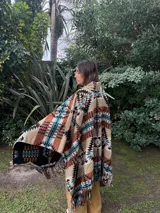 Poncho estilo navajo confeccionado en mezcla de lana y algodón, con estampado geométrico multicolor en tonos beige, marrón, negro, turquesa y blanco, y terminación de flecos en el ruedo.