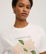 Remera de algodón color blanco, con cuello redondo y manga corta. Presenta un estampado con la frase 'Soy flor. Soy latina' y flores bordadas en relieve con textura tejida.