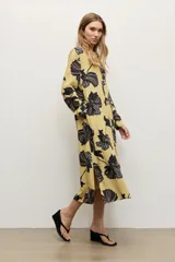 Vestido camisero midi de color amarillo maíz con estampado floral en tonos oscuros, de manga larga, cuello camisero, cierre frontal de botones y cinturón negro delgado con hebilla metálica.