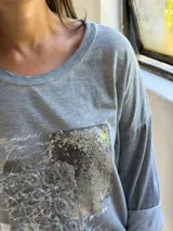 Remera gris de manga larga con cuello redondo y diseño holgado. Presenta un aplique frontal rectangular con detalles de lentejuelas plateadas y estampado tipográfico.