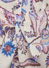 Vestido midi de algodón con estampado floral multicolor sobre fondo crudo. Presenta escote en pico, mangas largas con puños abotonados y un lazo decorativo en la cintura que crea un efecto drapeado, con una abertura frontal.