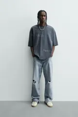 Camiseta de algodón color gris oscuro, corte relajado (relaxed fit), con cuello redondo y manga corta. Presenta dos inscripciones bordadas en hilo blanco en el frente.