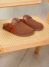 Zueco color marrón chocolate con diseño combinado de ecocuero liso en los laterales y gamuza sintética en la parte frontal. Cuenta con una tira con hebilla metálica ajustable en el empeine.