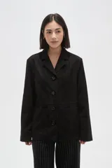 Chaqueta de gamuza sintética en color negro, con cuello solapa, cierre frontal con botones y bolsillos delanteros. Presenta un corte holgado y mangas largas.