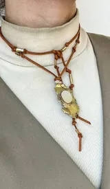 Gargantilla artesanal compuesta por un tiento de gamuza color tostado, decorado con tubos metálicos y un dije central de diseño irregular en metal dorado con una piedra central incrustada.