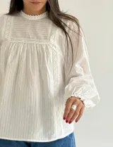 Blusa blanca de corte holgado con cuello redondo, mangas largas con puños elásticos y detalles de puntillas verticales en el frente.