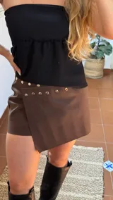 Pollera short de ecocuero color marrón, con diseño cruzado y detalle de tachas metálicas en la parte frontal. Cuenta con cierre ajustable mediante botones.