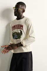 Camiseta de manga larga y cuello redondo en color crudo, con estampado gráfico frontal de paisaje montañoso y tipografía en tonos granate y verde.